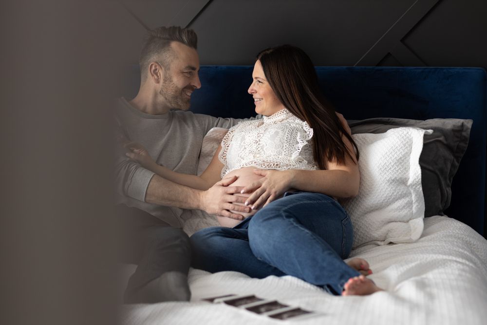 Smiling pregnant woman sitting on bed with partner, both touching her baby bump, with ultrasound photos nearby.