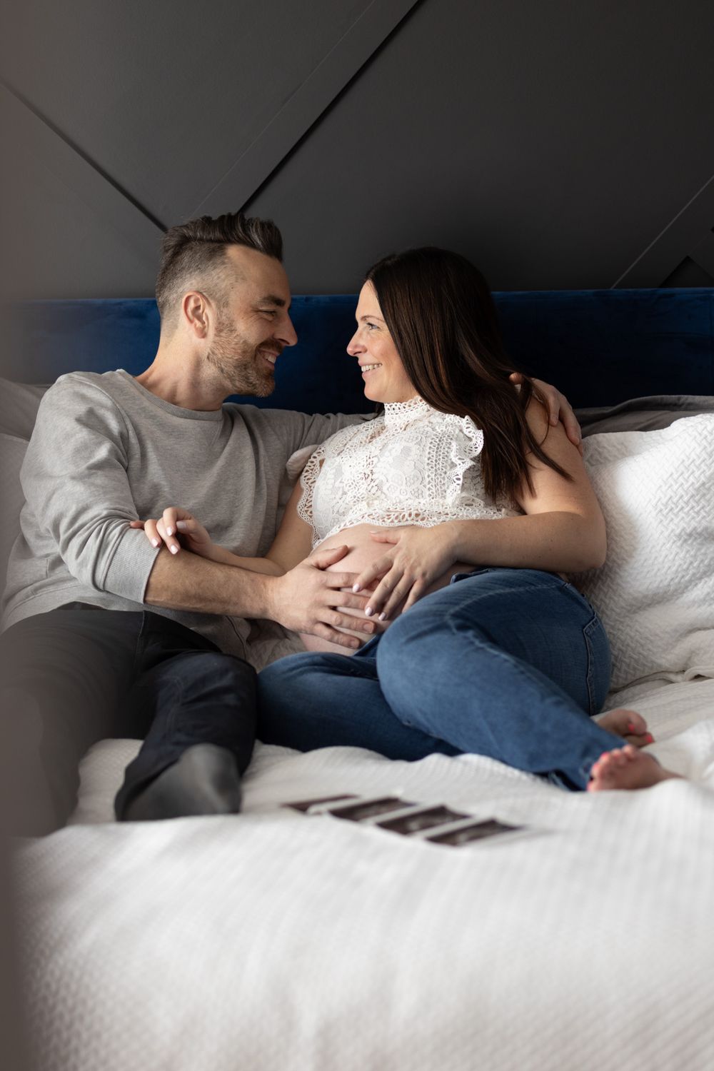 Happy couple sitting on bed, smiling at each other as man tenderly touches pregnant woman's baby bump.