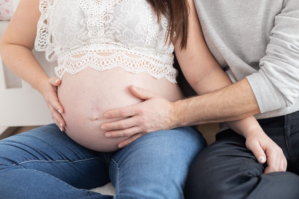 Pregnant woman in jeans with partner's hand gently resting on her bare baby bump during maternity photo session.