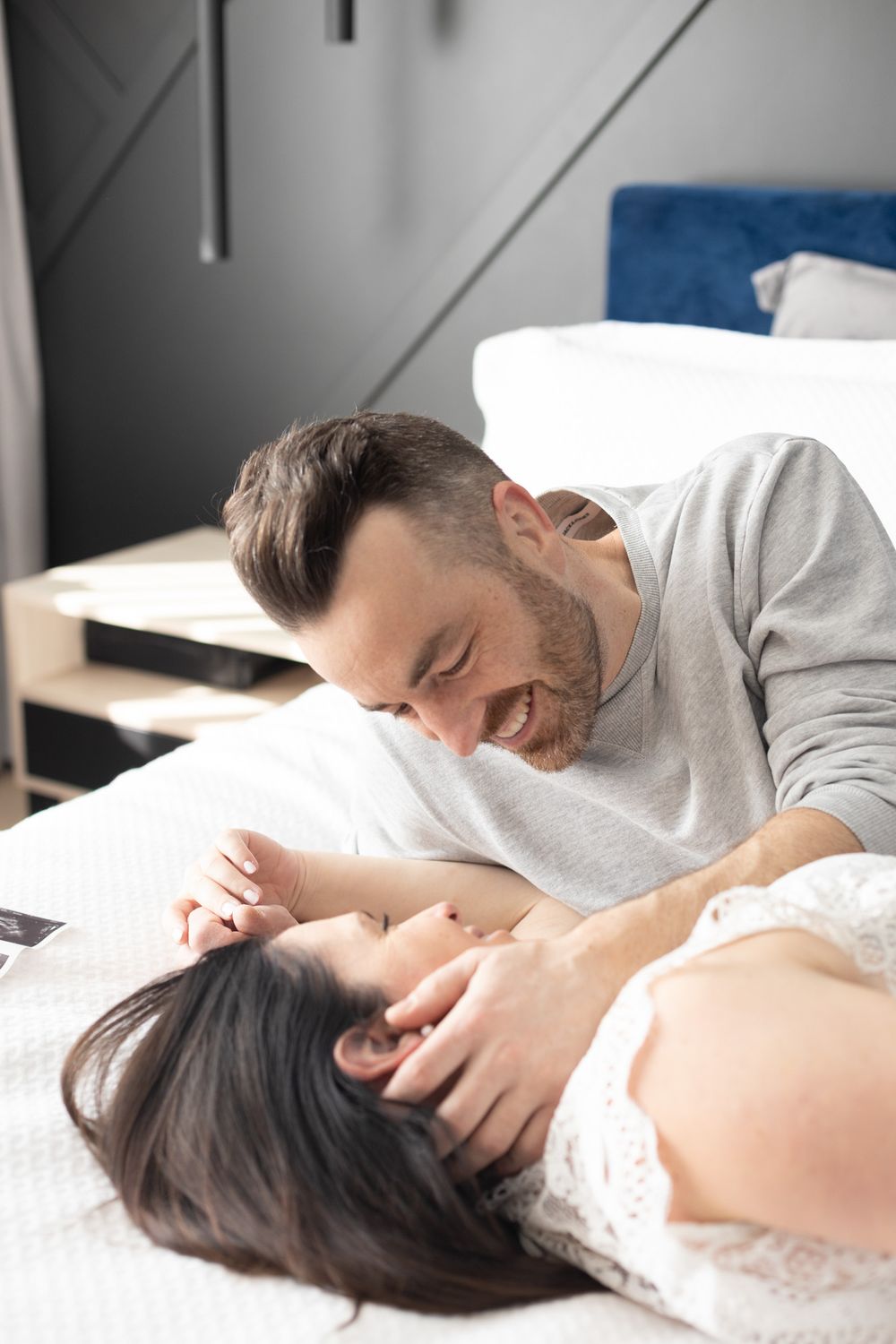 Happy couple laughing and bonding together while lying on a white bed in a modern bedroom.