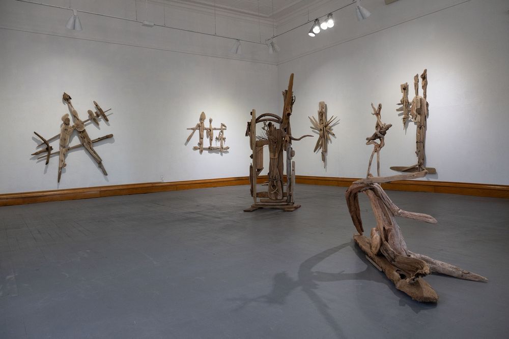 Gallery installation featuring abstract driftwood sculptures arranged on gray floor and mounted on white walls.