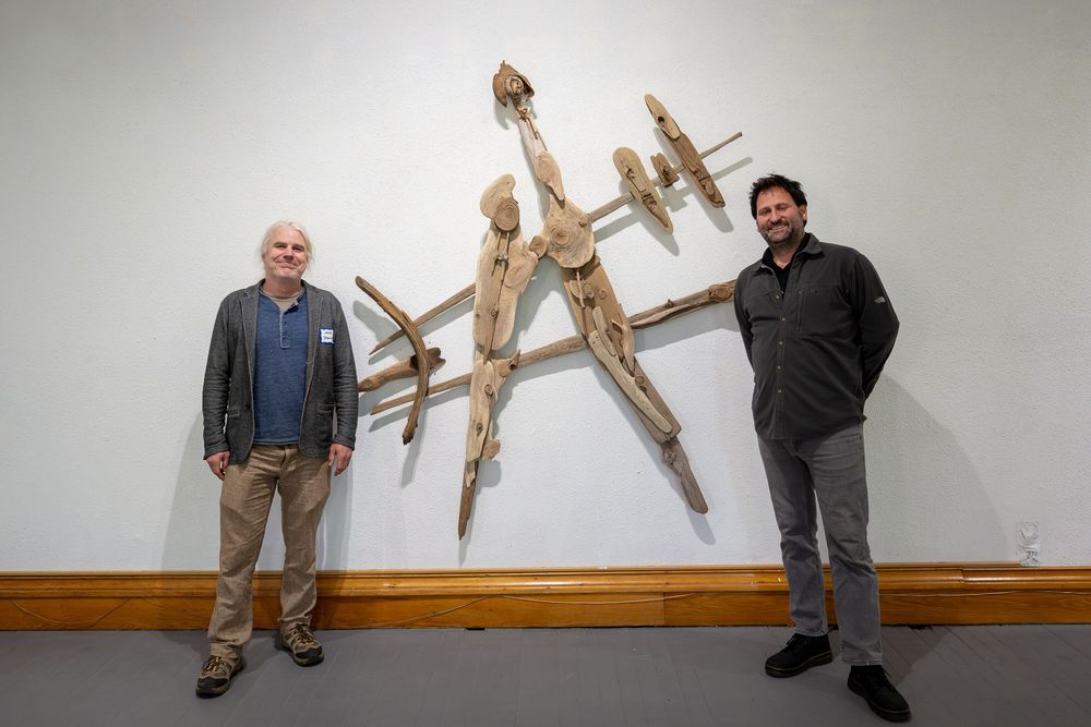 Two men stand beside a large wooden sculptural wall installation in an art gallery.