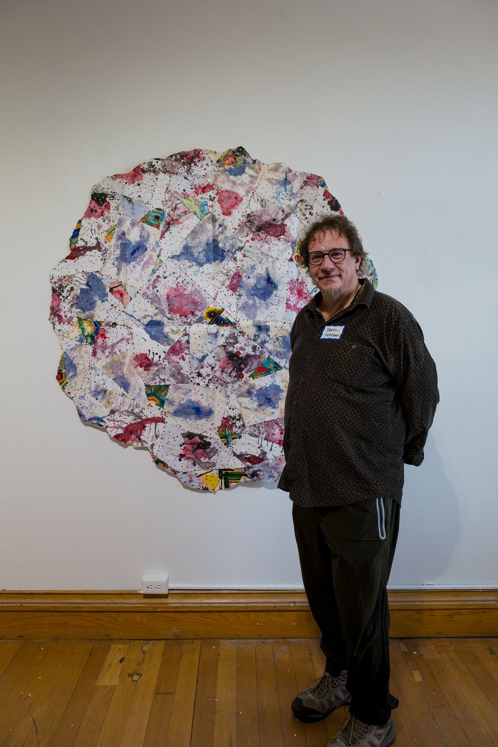 Man standing beside large colorful abstract circular artwork on white gallery wall with wooden floor.
