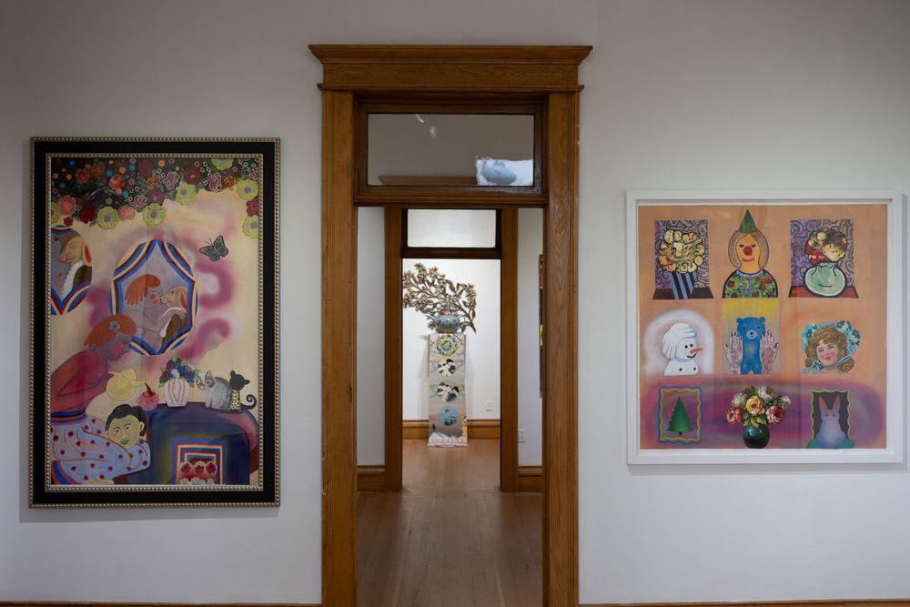 Art gallery interior with colorful paintings on white walls flanking a doorway leading to a sculpture in the next room.