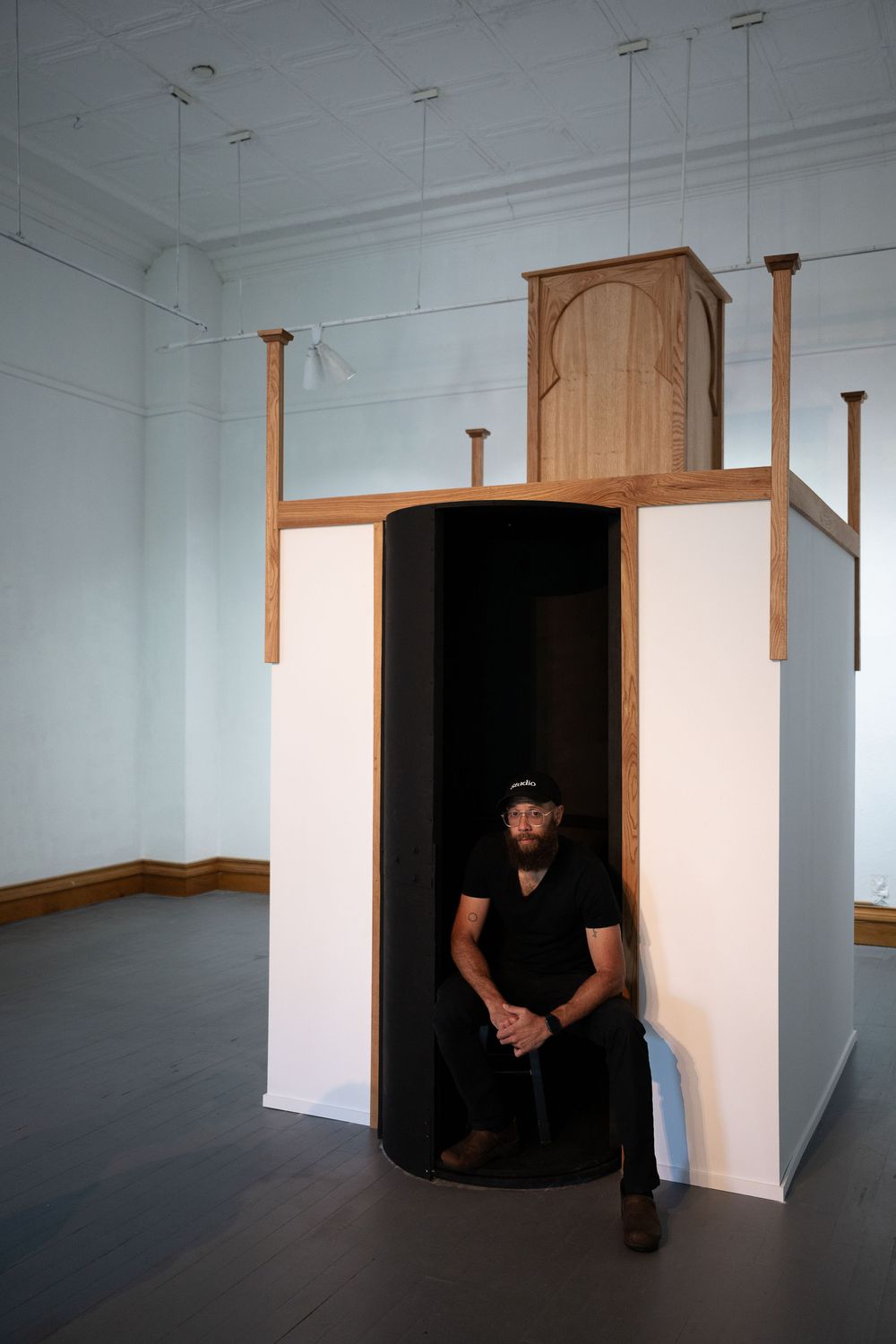 Man in black outfit sits in curved dark booth beneath ornate wooden minbar structure in white gallery space.