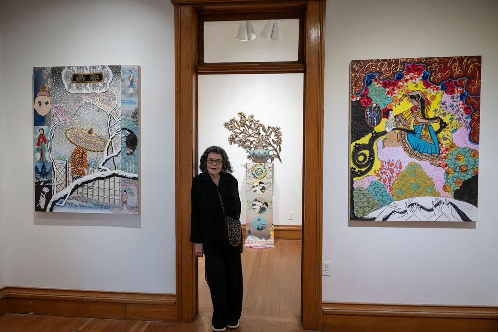 Woman in black standing between colorful contemporary paintings in a white gallery exhibition space.