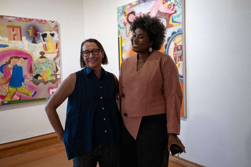 Two women pose in an art gallery surrounded by colorful contemporary paintings on white walls.