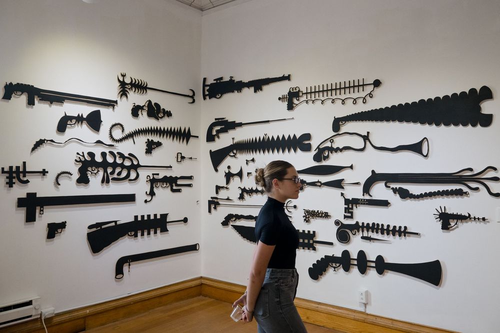 Woman walks past a wall display of black sculptural gun-shaped artworks in a white gallery with hardwood floors.