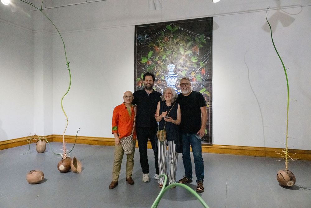 Four people pose in an art gallery with tall plant sculptures and a large colorful painting on the wall.