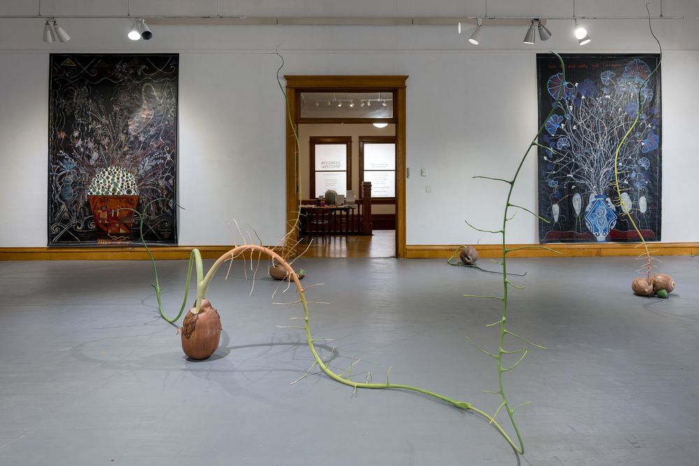 Gallery installation with dark botanical paintings on white walls and sculptural vine-like forms with ceramic vessels on gray floor.