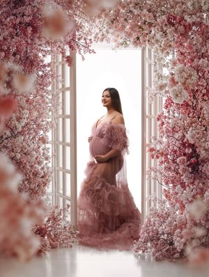 A dreamy maternity portrait shows an expectant mother in a pink ruffled gown framed by cherry blossoms and white doors.