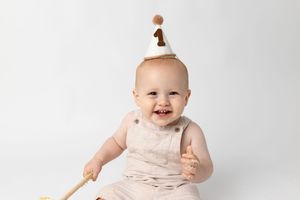 Smiling baby in a tan romper and a white party hat with the number one sits on a white background, celebrating a first birthday.