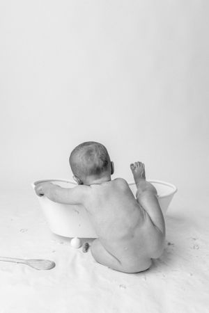 Black and white photo of a baby seen from behind leaning into a small white tub, with a wooden spoon and egg nearby on a soft backdrop.