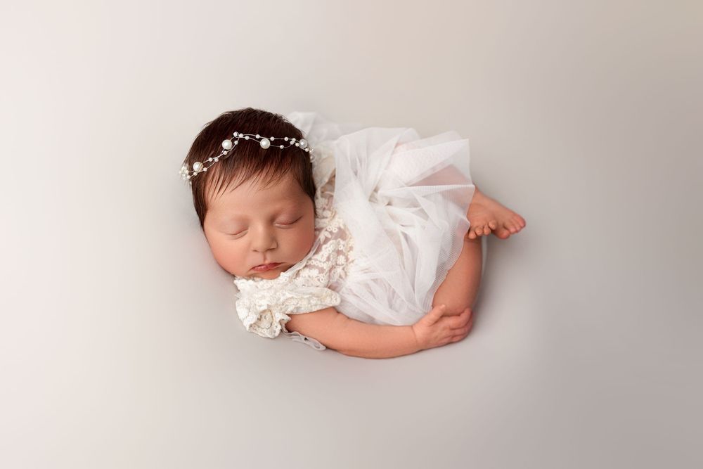 A sleeping newborn baby wearing a delicate white lace dress and pearl headband rests peacefully on a light gray backdrop.