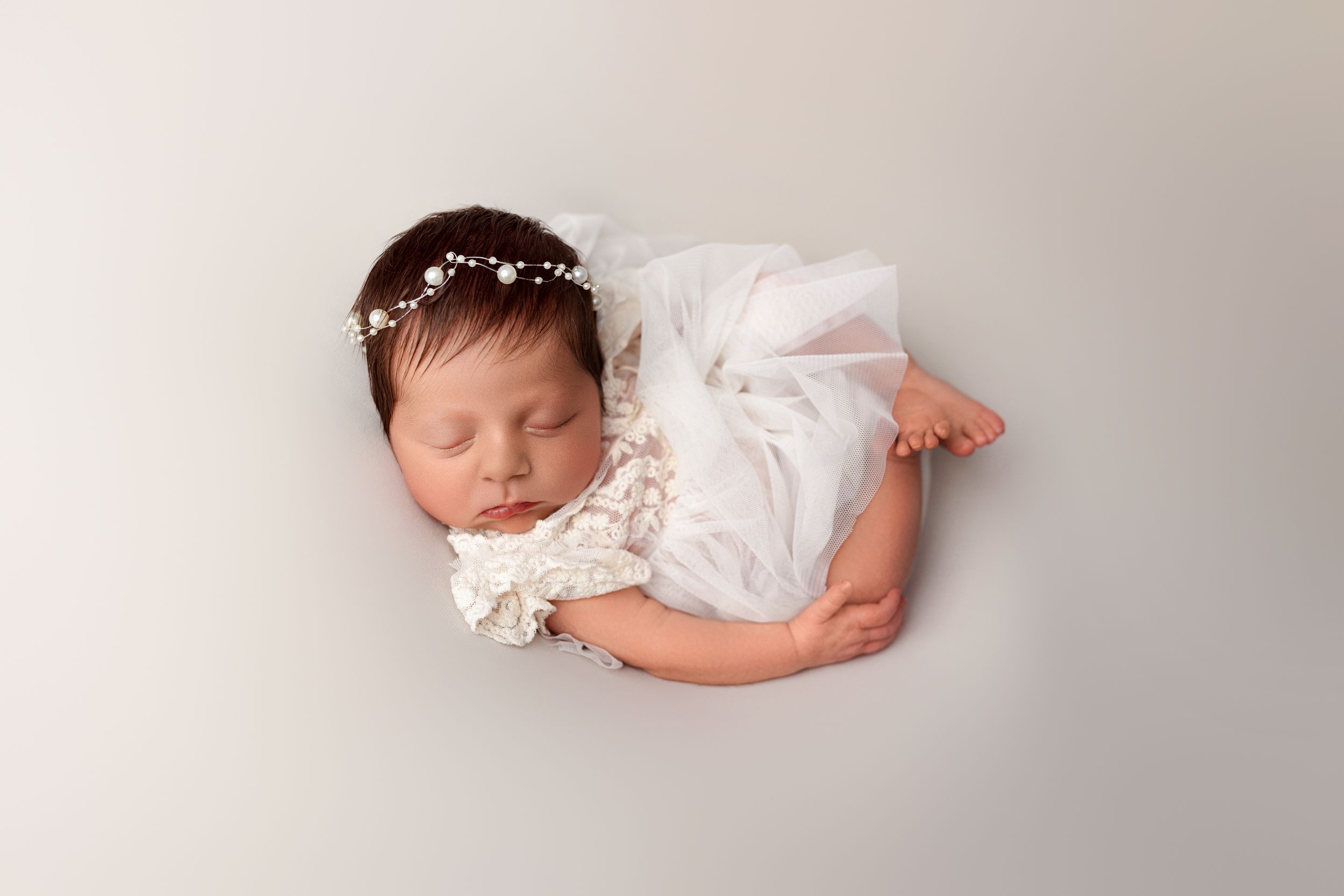 A sleeping newborn baby wearing a delicate white lace dress and pearl headband rests peacefully on a light gray backdrop.