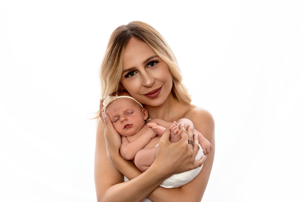 Smiling blonde woman cradling a sleeping newborn baby against her chest in a bright, minimalist newborn portrait on a white background.