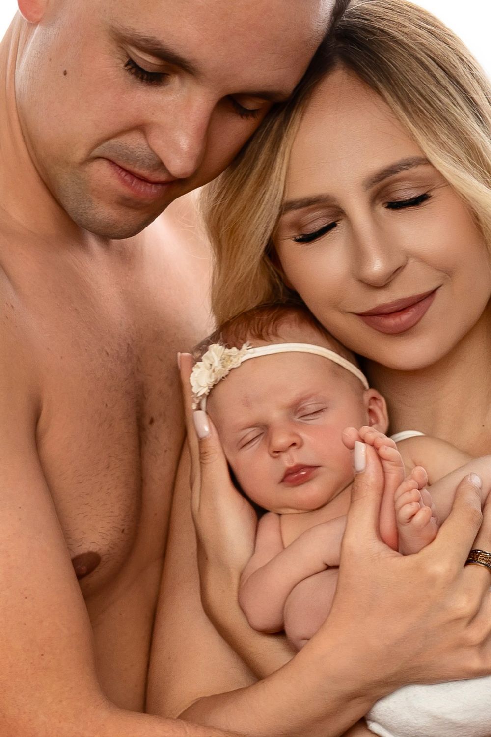 Smiling parents with closed eyes gently cradle their sleeping newborn baby wearing a floral headband in a warm, intimate family portrait.