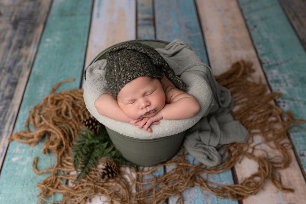 Sleeping newborn in pale gray metal bucket on rustic turquoise wooden backdrop with gauze and jute decorations.