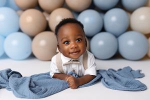Baby in white collared shirt sits with denim fabric against blue and tan balloon wall.