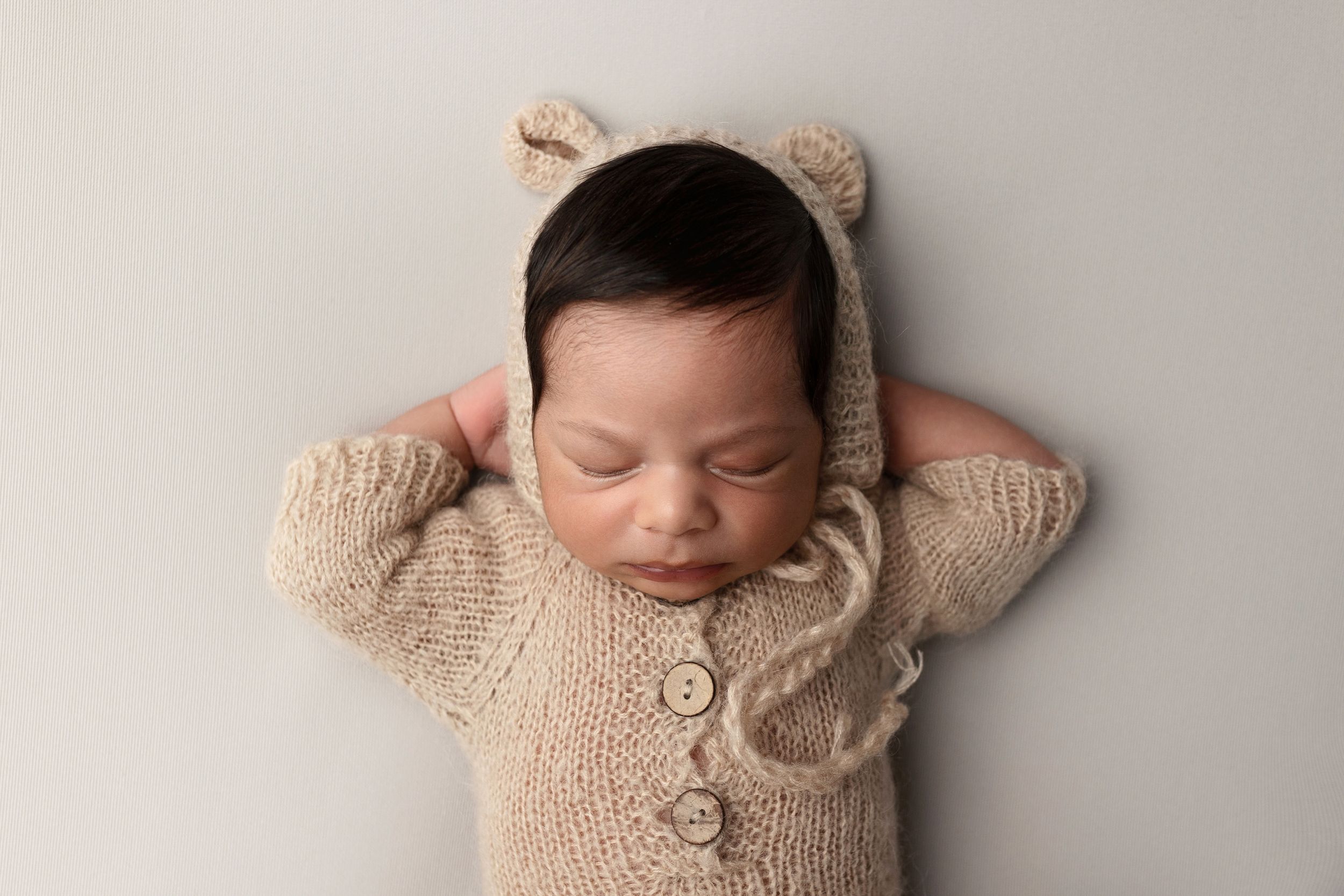 Sleeping newborn baby in a beige knit bear bonnet and button sweater poses with hands behind head on a soft neutral backdrop.