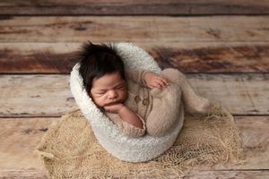 Sleeping newborn baby with dark hair in a cream knit outfit curled up in a cozy white bowl on burlap over a rustic wooden floor.