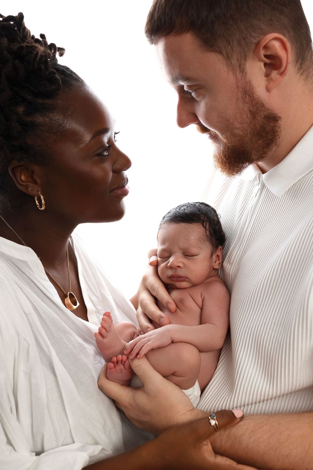 Smiling parents in white shirts gaze lovingly at each other while cradling their sleeping newborn baby in a bright newborn family portrait.
