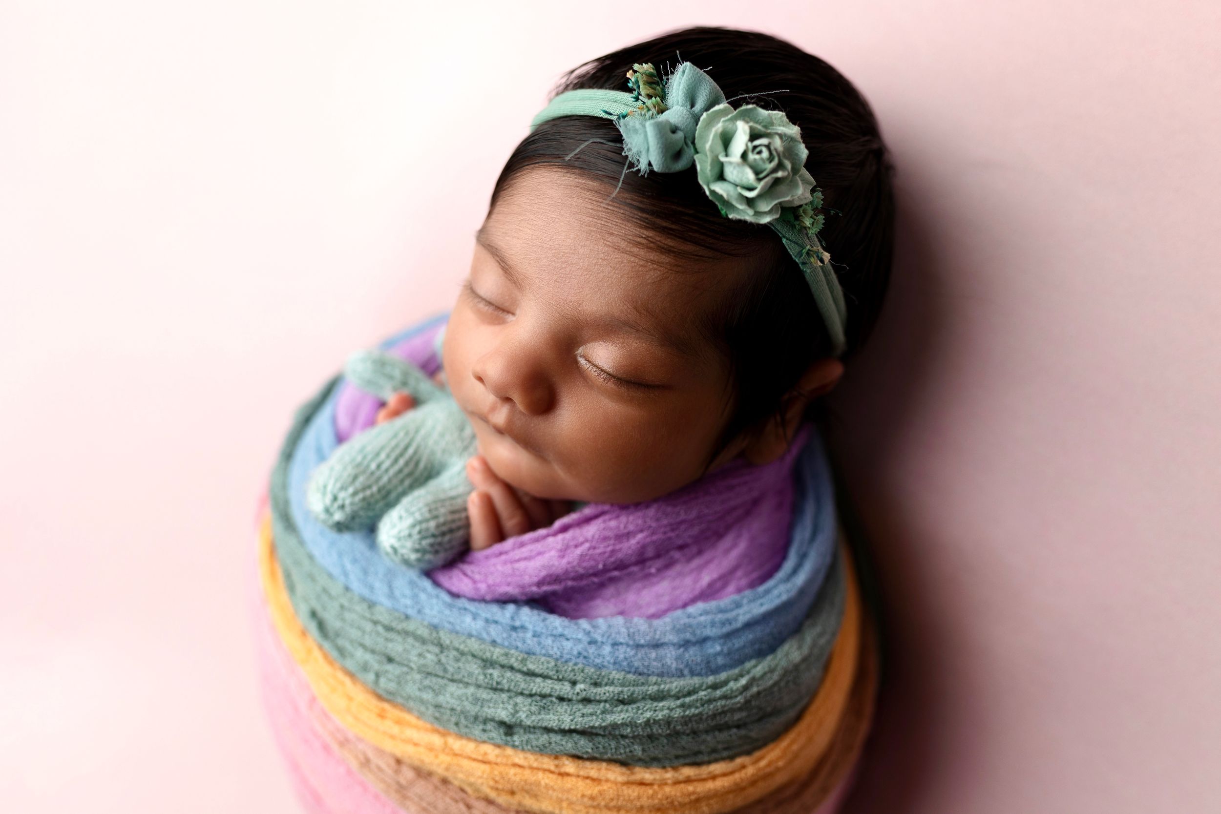 Sleeping newborn baby wrapped in a pastel rainbow swaddle wearing a teal flower headband on a soft pink backdrop.