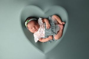 Sleeping newborn photographed through heart-shaped frame wearing white and gray outfit.