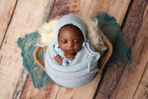 Sleeping newborn wrapped in soft blue swaddle while resting in a vintage teacup on rustic wooden floor.