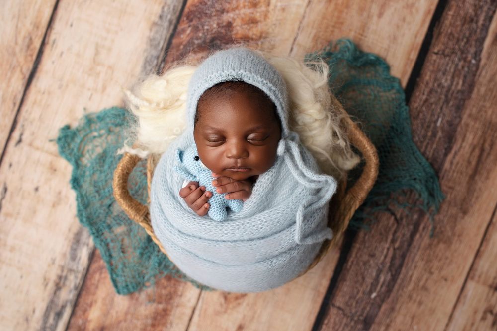 Sleeping newborn wrapped in soft blue swaddle while resting in a vintage teacup on rustic wooden floor.
