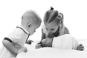 Black and white photo of two young siblings leaning over to gently kiss and admire a sleeping newborn baby wrapped in a soft blanket.