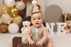 Baby in birthday hat sitting on wooden box with gold and white balloon backdrop.