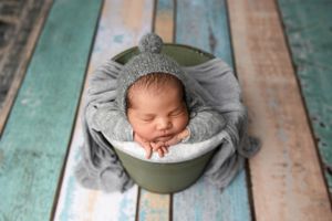 Sleeping newborn in grey bonnet resting in circular bowl prop on turquoise wooden floor.