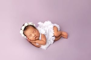 A sleeping newborn baby wearing white clothing lies on lavender fabric in a professional photography setting.