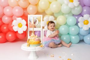 Baby celebrating with yellow cake and rainbow balloon arch during first birthday photoshoot.