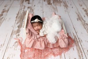 A newborn in a pink lace dress sleeps peacefully on a rustic wooden floor with a white plush toy.