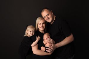 Portrait of a family dressed in black against dark background with toddler and newborn baby posing in studio setting.
