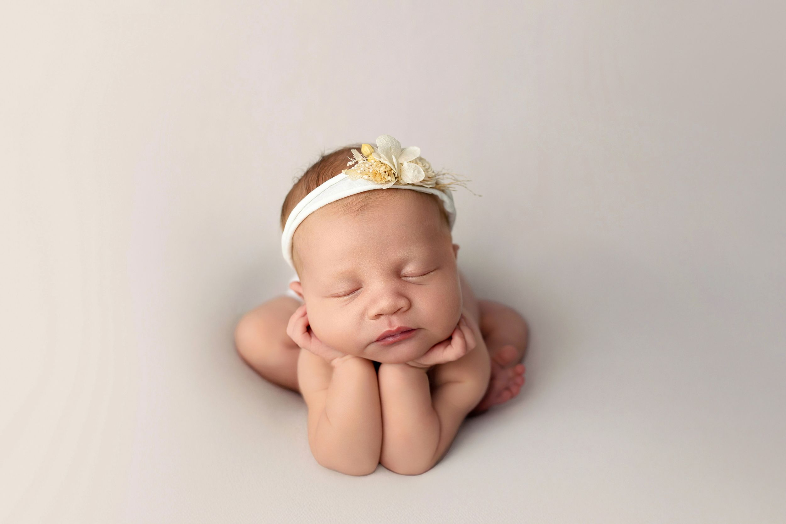 Sleeping newborn baby wearing a delicate crown headband poses with hands under chin on cream backdrop.