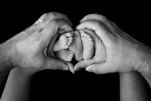 Adult hands forming a heart shape around tiny baby feet in black and white photography.