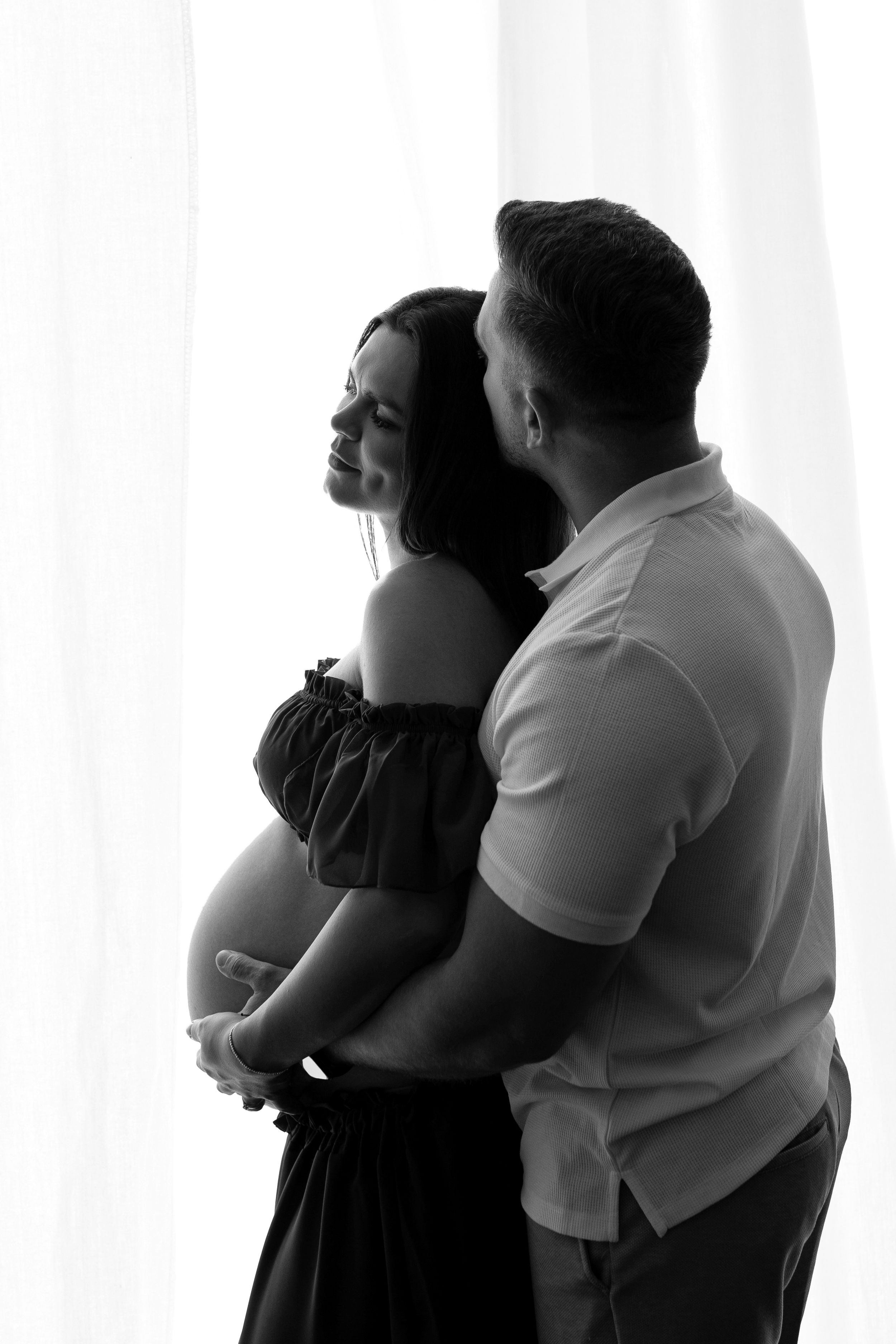 Black and white maternity portrait of a man embracing his pregnant partner from behind, both framed by soft sheer curtains.