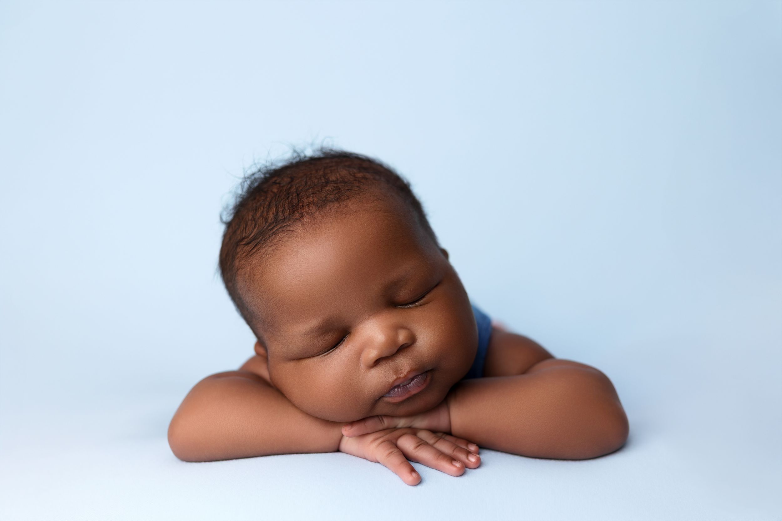 Peaceful sleeping baby laying on crossed arms against light blue background in professional portrait setting.