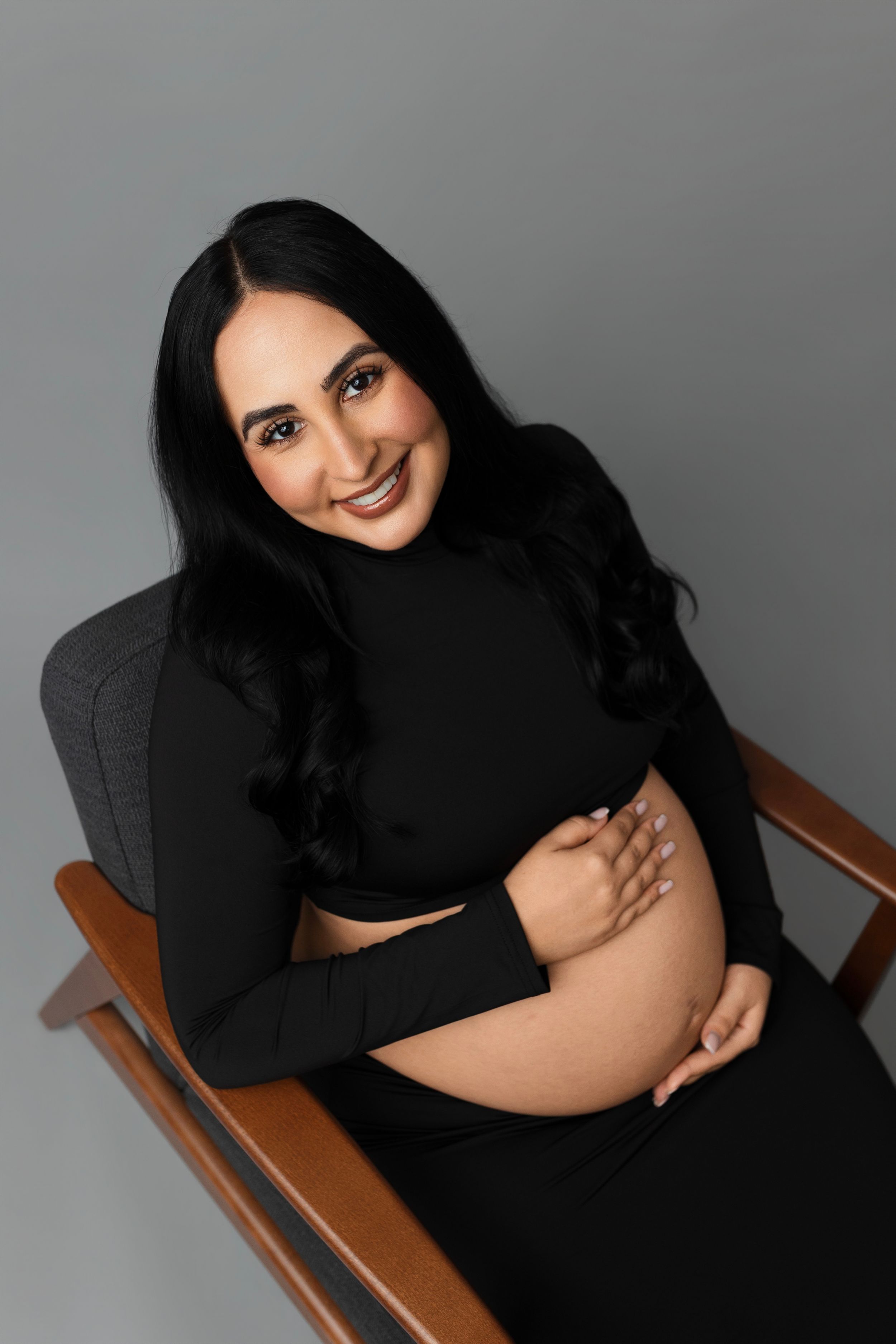 Smiling pregnant woman with long dark hair in a black crop top and skirt cradles her bare belly while seated in a mid-century wooden chair.