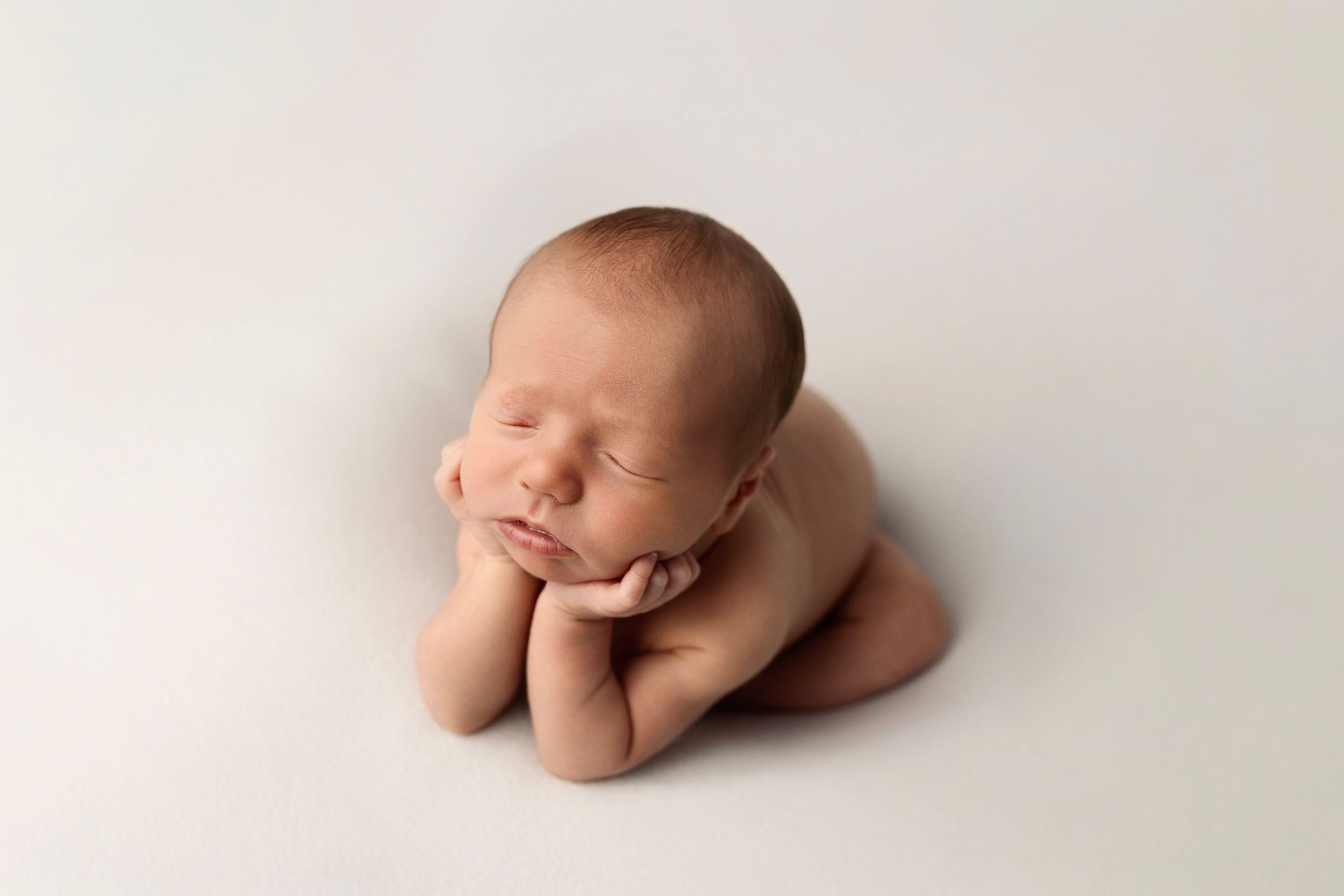 Sleeping newborn baby posed in froggy pose with chin resting on hands against a soft cream backdrop in a newborn photography session.
