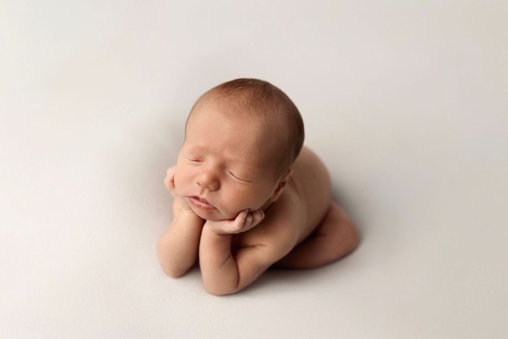 Sleeping newborn baby posed in froggy pose with chin resting on hands against a soft cream backdrop in a newborn photography session.