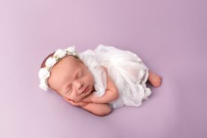 Sleeping newborn in white dress and floral headband on purple backdrop during professional photo shoot.