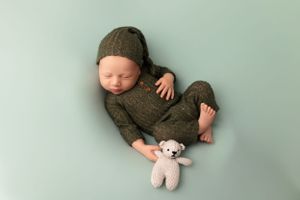 Sleeping newborn in knit outfit lying next to small teddy bear on mint green background.