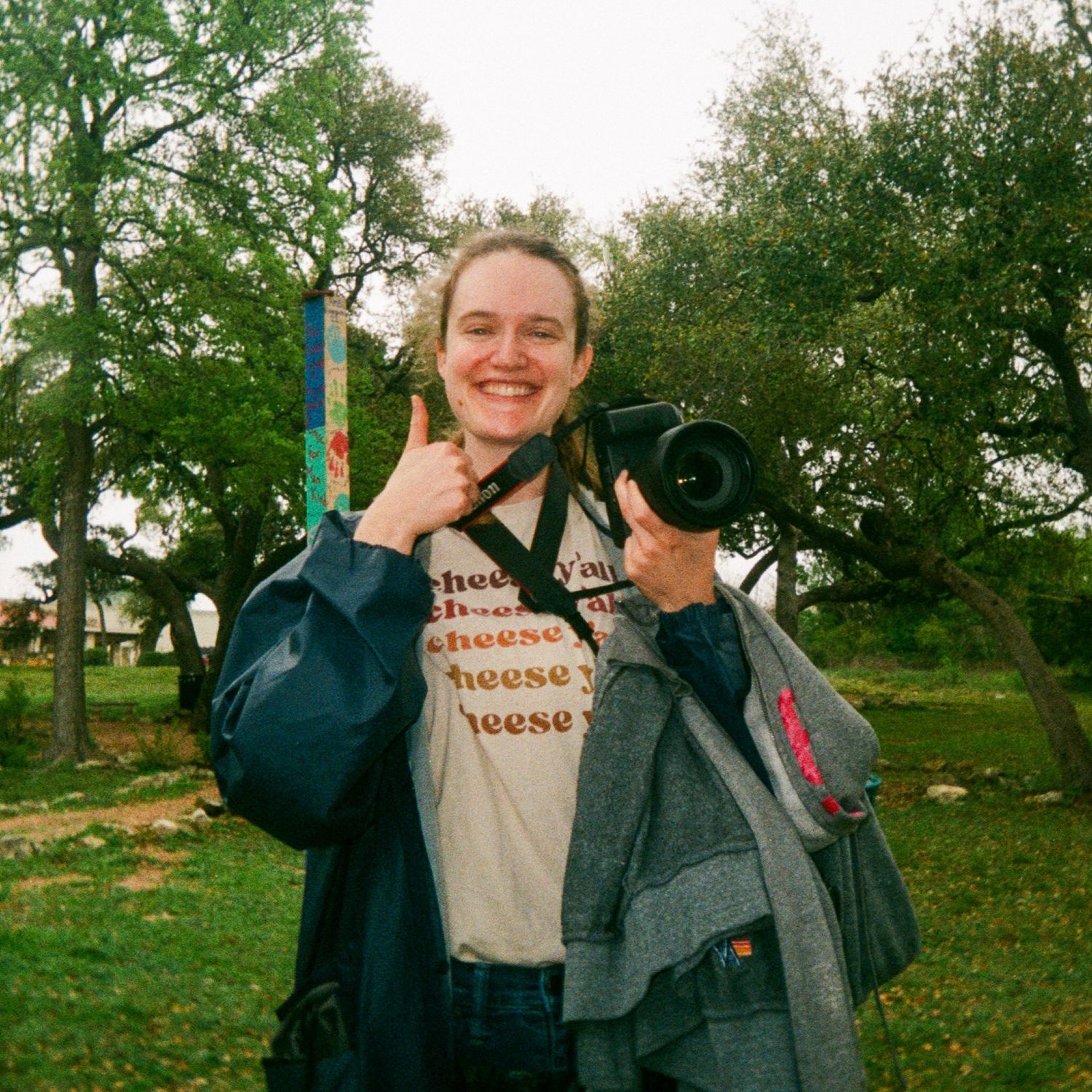 Photographer showing thumbs up sign while holding camera equipment in park setting.