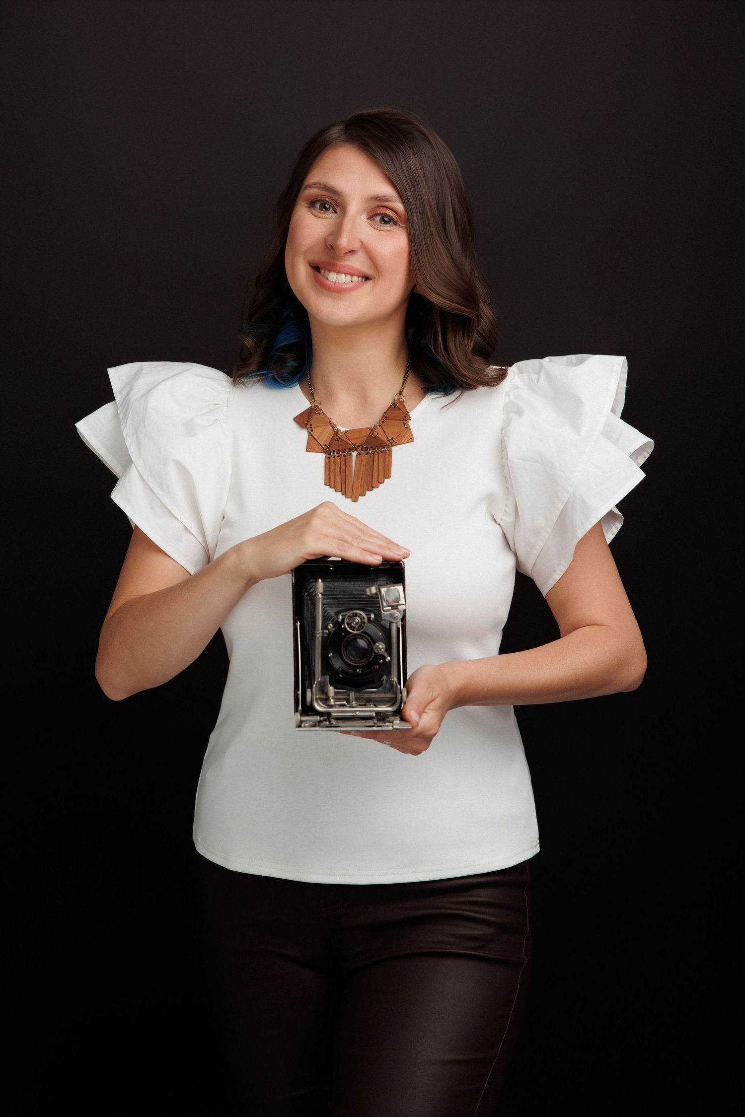 Small child in white lace dress holding wooden toy camera against white background.
