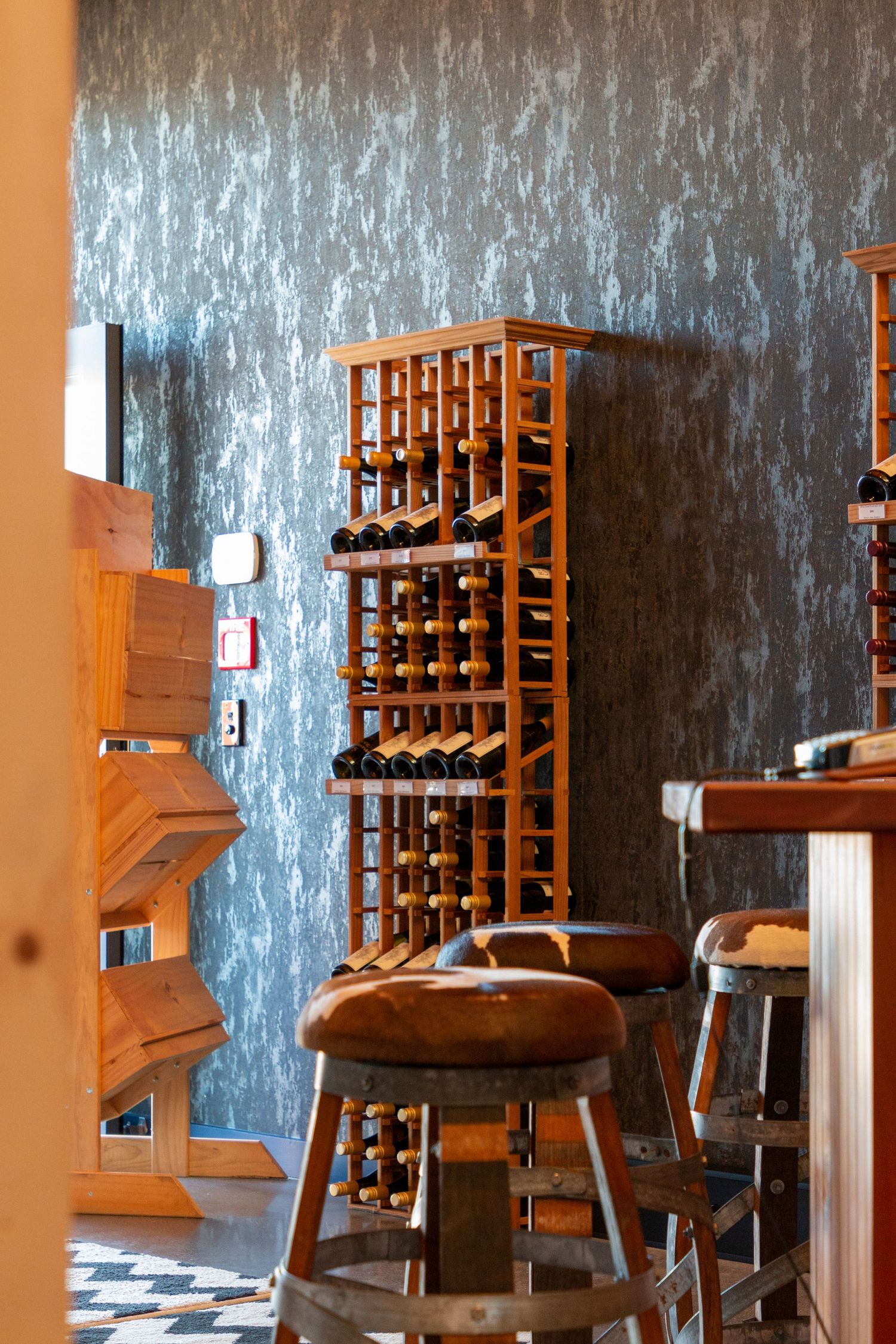 Warm interior with wooden bar stools, a wine rack filled with bottles, textured wallpaper, and soft natural lighting.