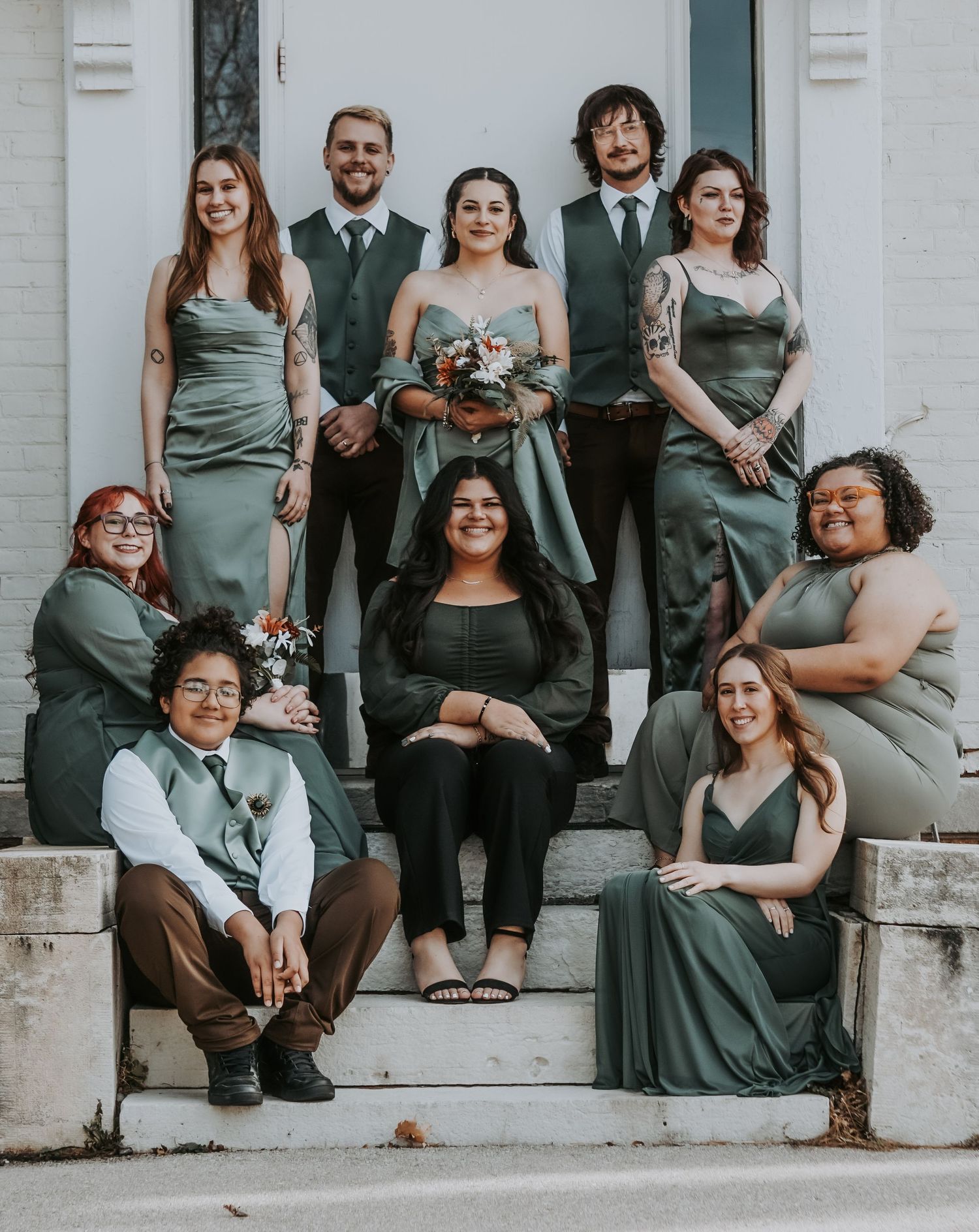 Wedding party in sage green attire poses casually on white building steps.
